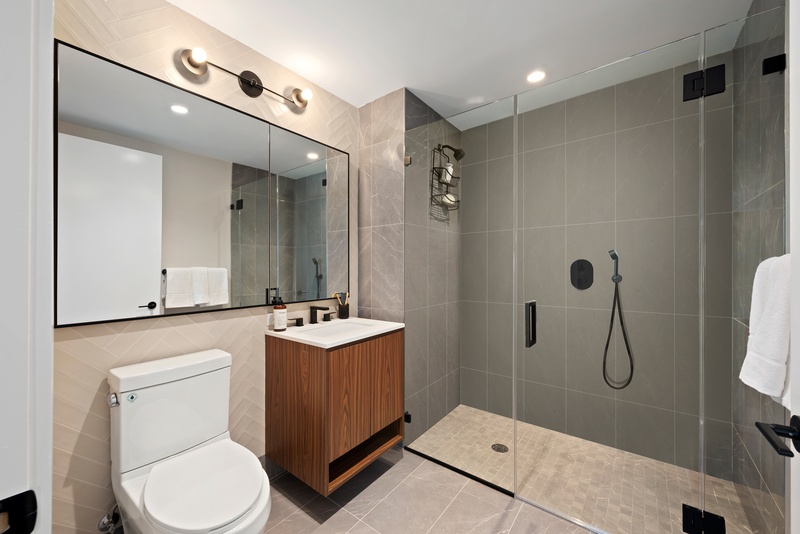 Modern bathroom with light and grey tiling throughout including walls, floors and large standing shoert, large window above both toilet and wood vanity with white sink