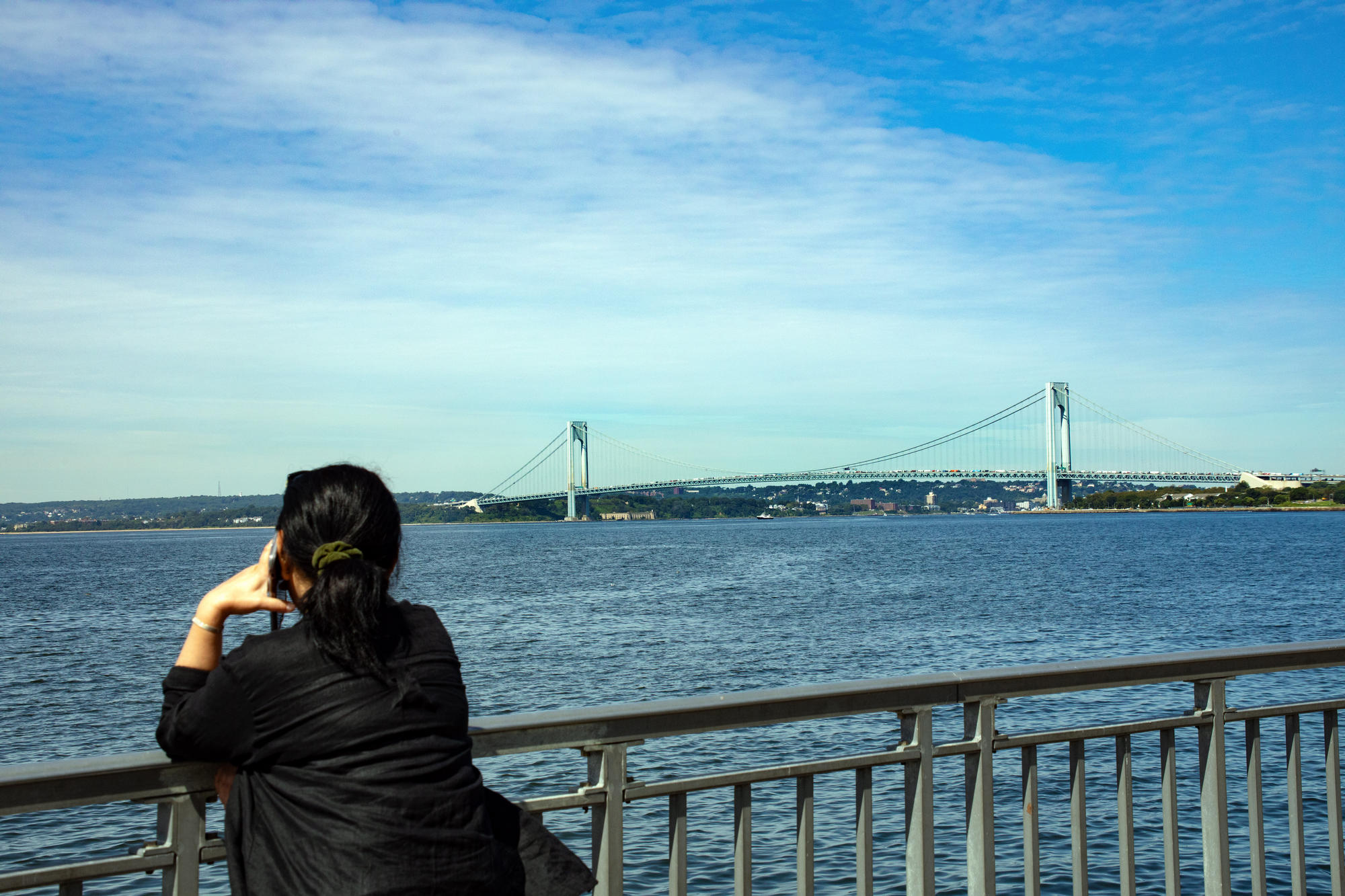 Woman with dark hair ponytail on phone overlooking water and view of bridge