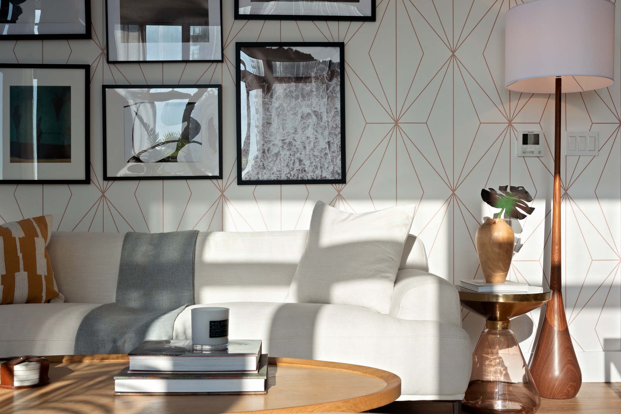 Detail view of white couch in living room with coffee table and books, candle, plants and lamp on side, throw pillows, grey throw blanket, wallpaper accent wall with framed photography