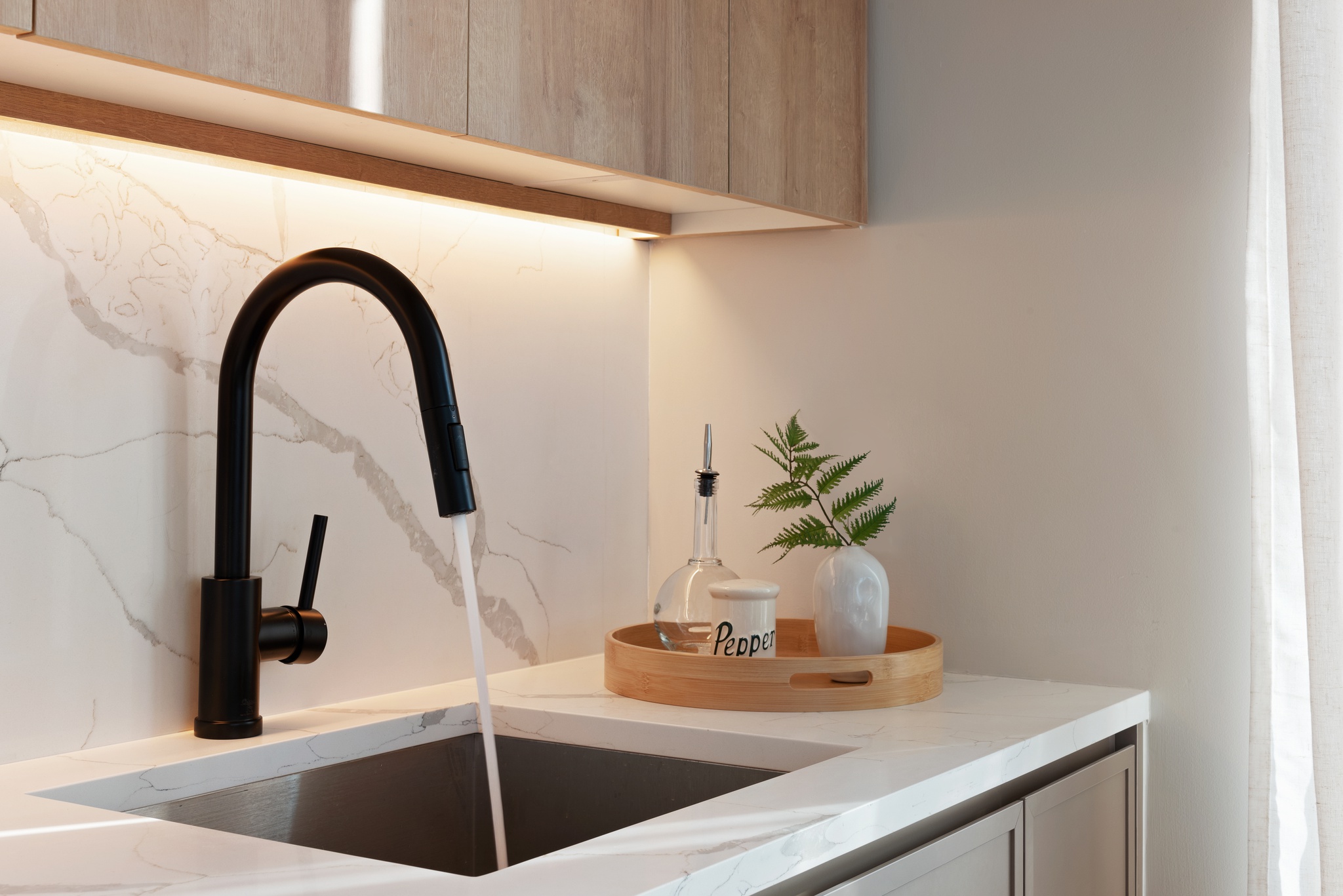 Detail of black kitchen sink faucet with white marble looking counter and backsplash, white walls, and light wood cabinetry