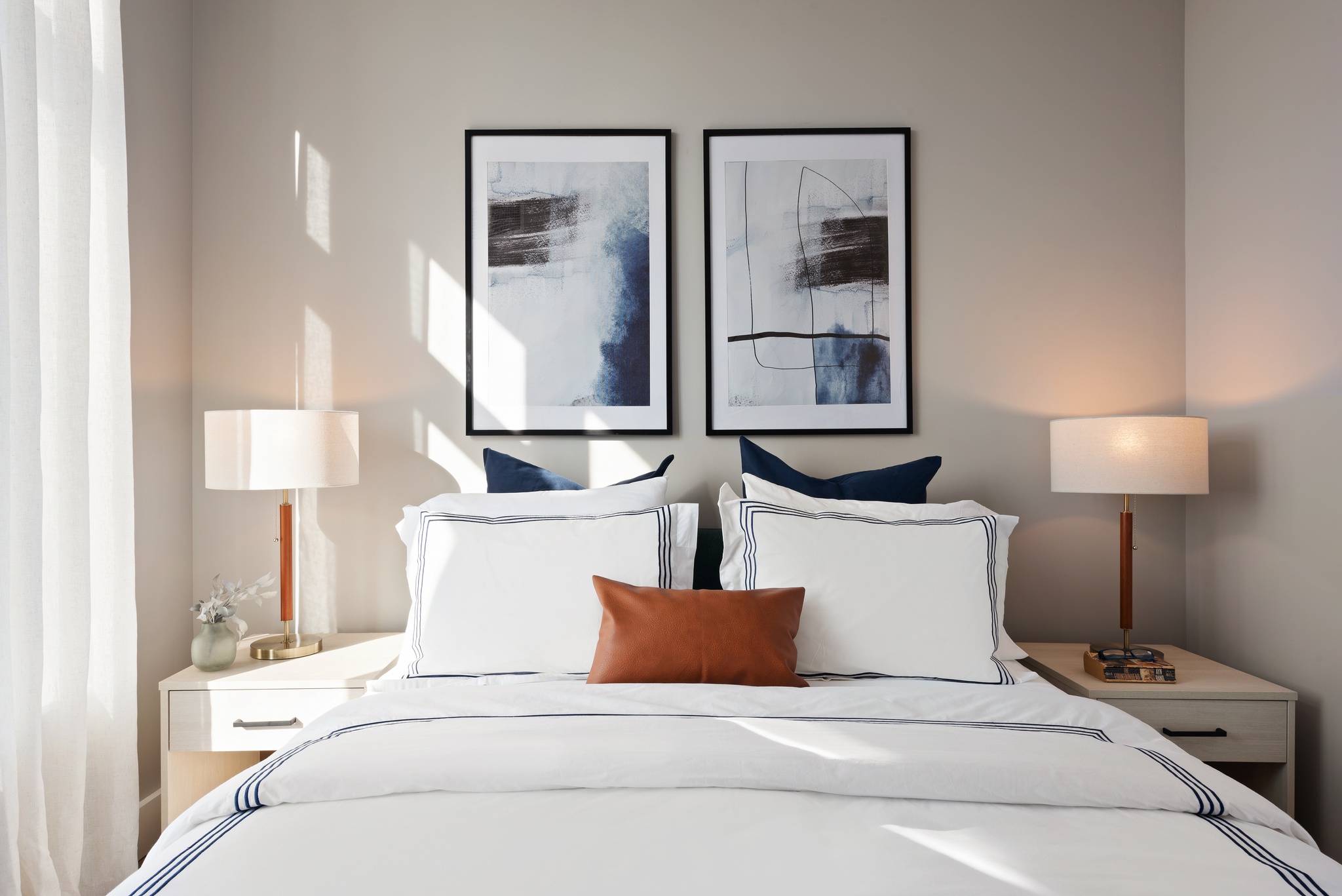 detail of bed with navy and white bedding, white nightstands on sides with drawer and lamps, 2 large square modern framed art behind bed on light grayish wall