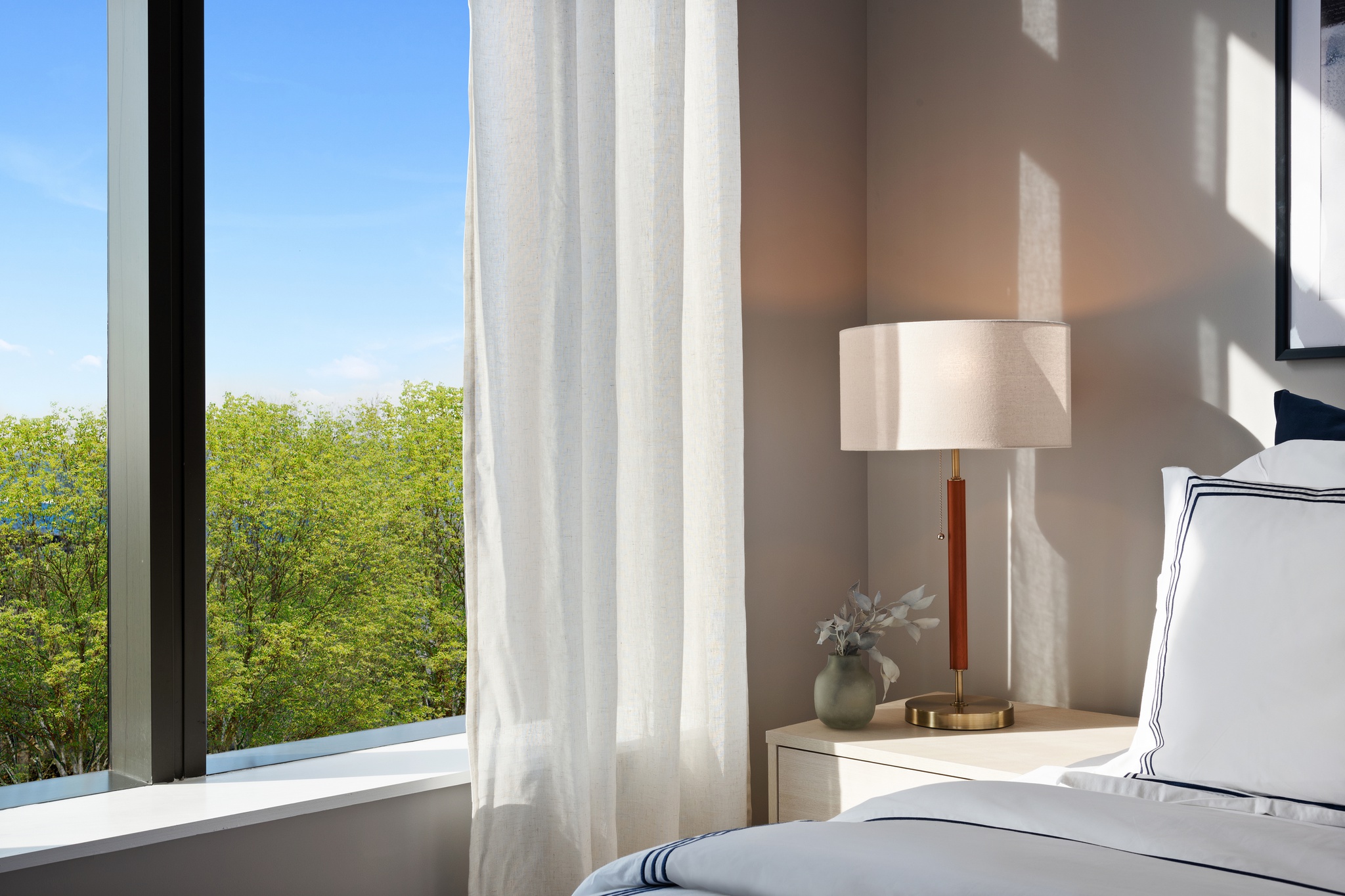 Corner shot of bed with white and navy bedding, nightstand with lamp and window with white curtains