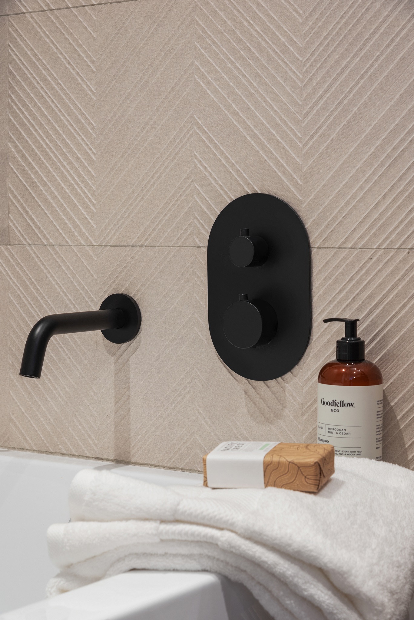 black bath faucet fixtures, shampoo bottle and textured light colored tiles on walls