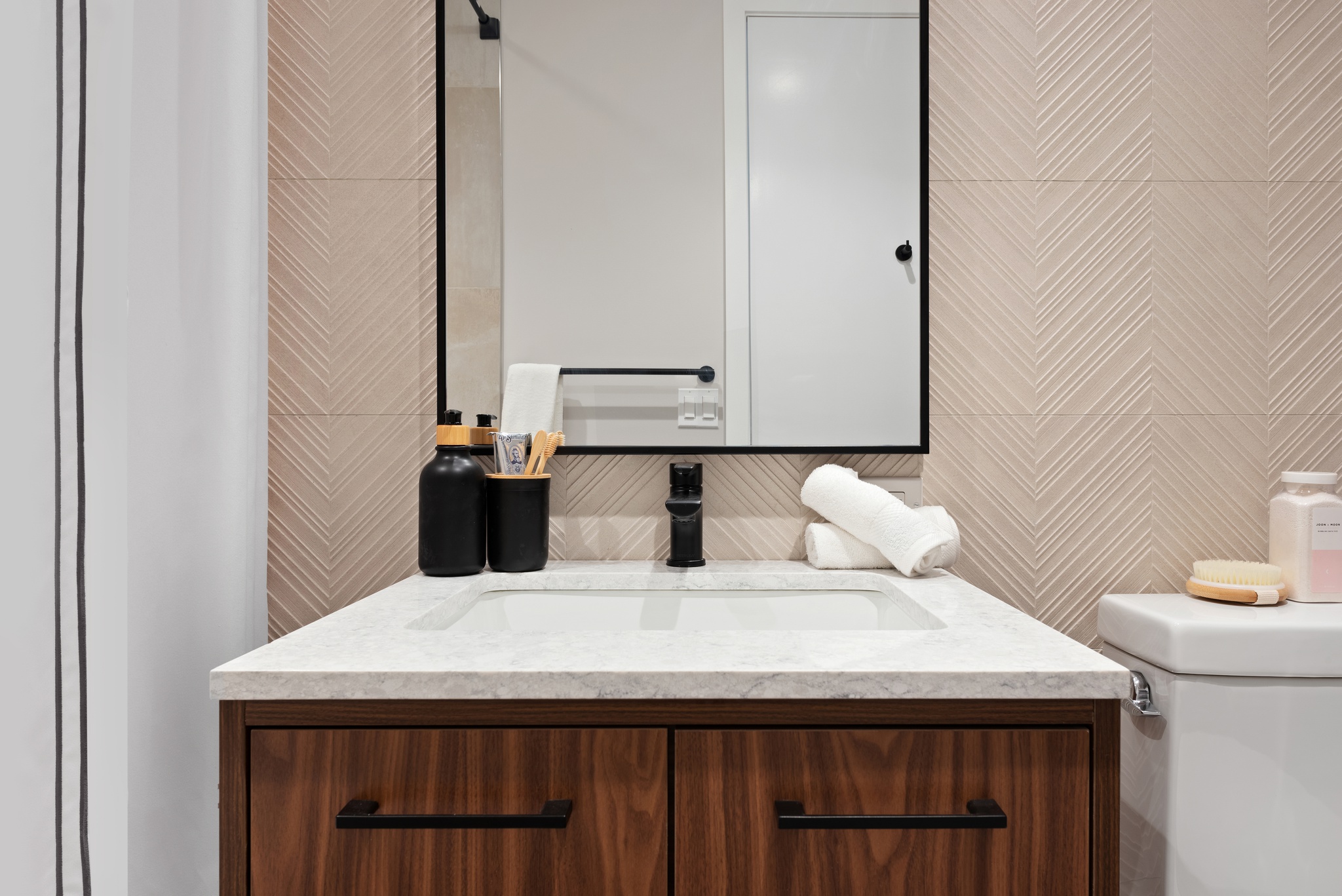 wood vanity detail with black faucet, cup and dispenser, and white sink and vertical rectangle mirror above, textured light colored tiles on walls