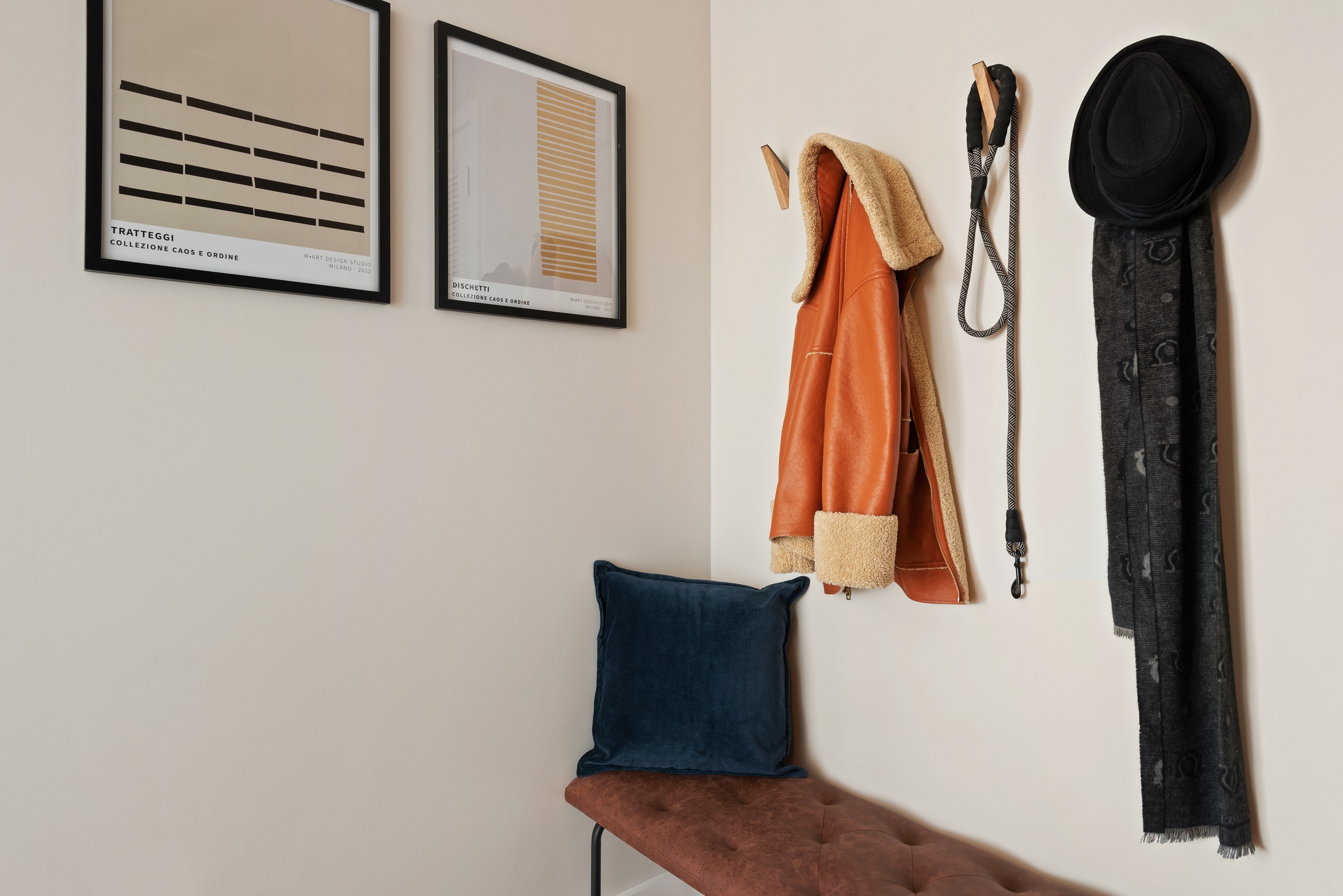 corner wall shot showing detail of wood wall hooks with coat, dog leash, hat and scarf hanging above bend with navy pillow and 2 square framed art pieces on wall next to it