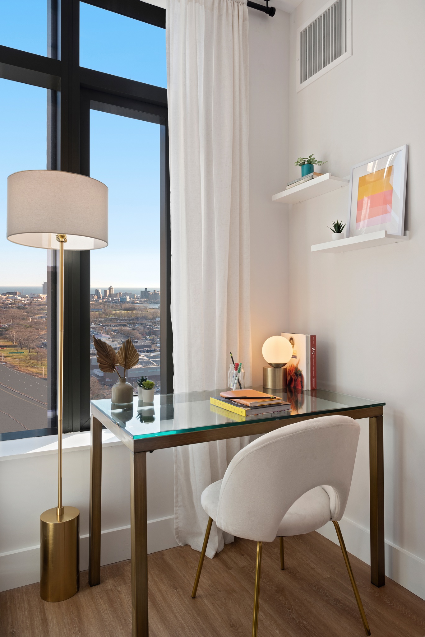 Detail of glass desk in bedroom with books and supplies, floor lamp and shelves on side, all in front of window