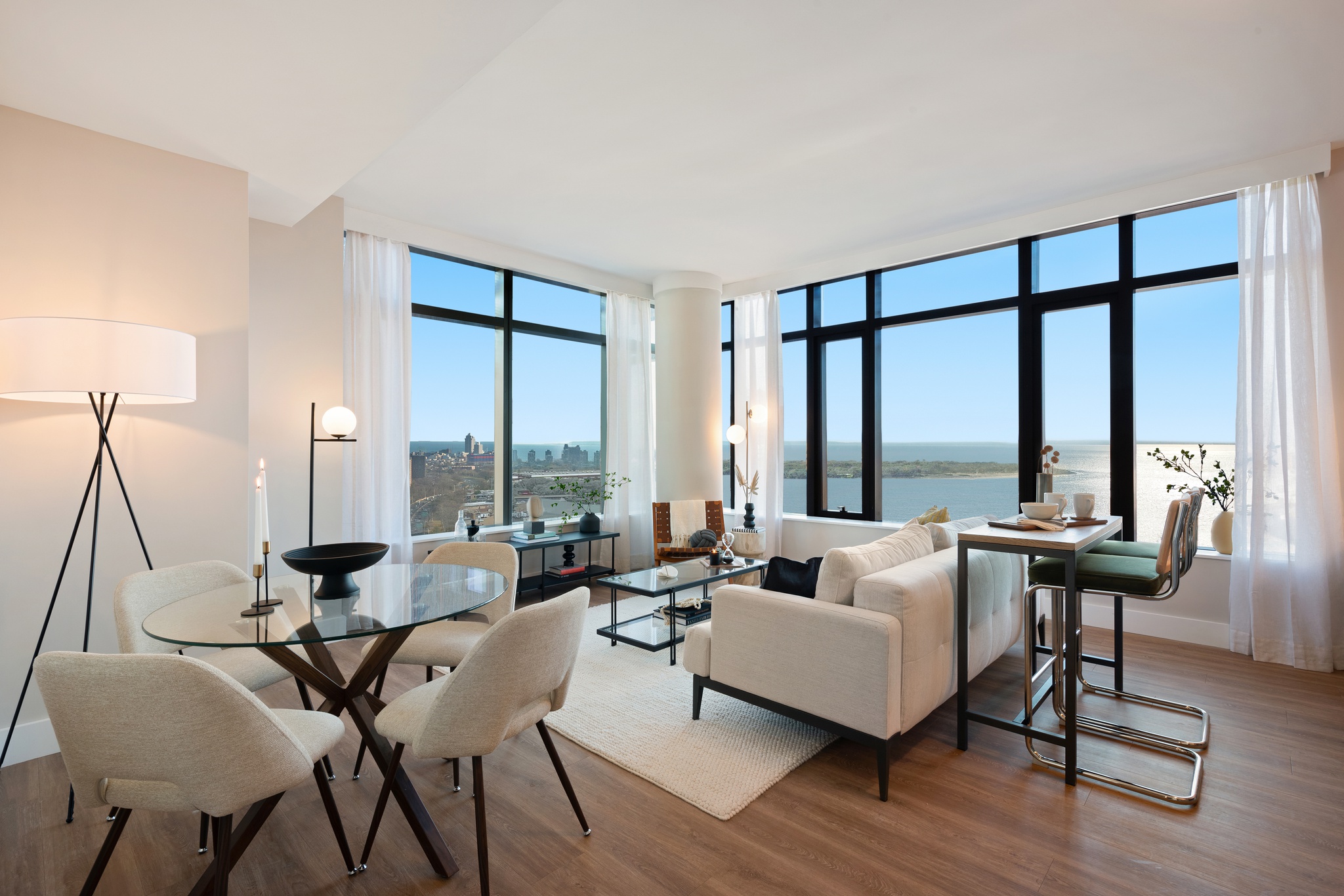 view of bright, modern living room with white couch, glass round dining table and seating for 4, area rug on wood floor, modern lamps, side table with high stool chairs, large windows around corner with white curtains