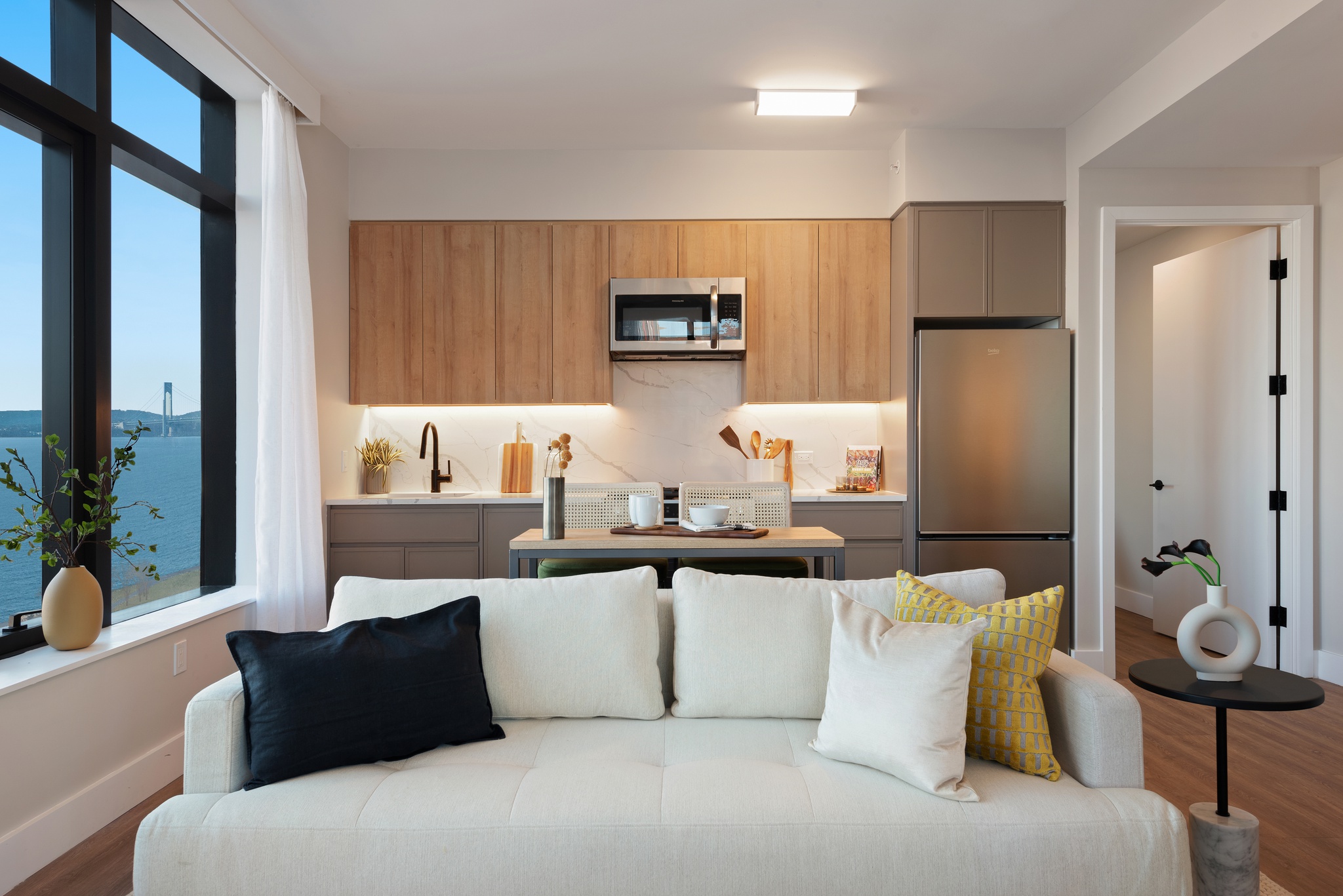 view of bright kitchen with light wood and greyish cabinetry, stainless steel appliances behind white couch in living area an window on the side with water views