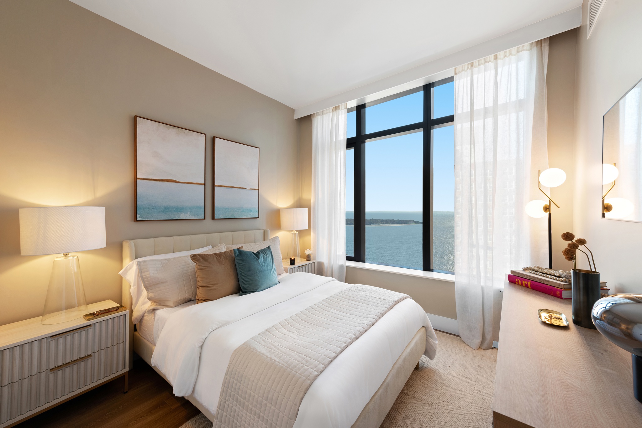 bright modern bedroom with rug on wood floor, white 2 drawer night stands on sides with modern lamps, wood dresser with lamp, books and vase and rectangular mirror on wall above it across from bed, large window with white curtain and floor lamp next to it, 2 large square art pieces behind and above bed on light greyish wall